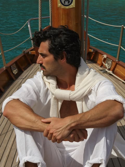 Man in ivory linen shirt with knit tied at the neck seated on a sailboat deck