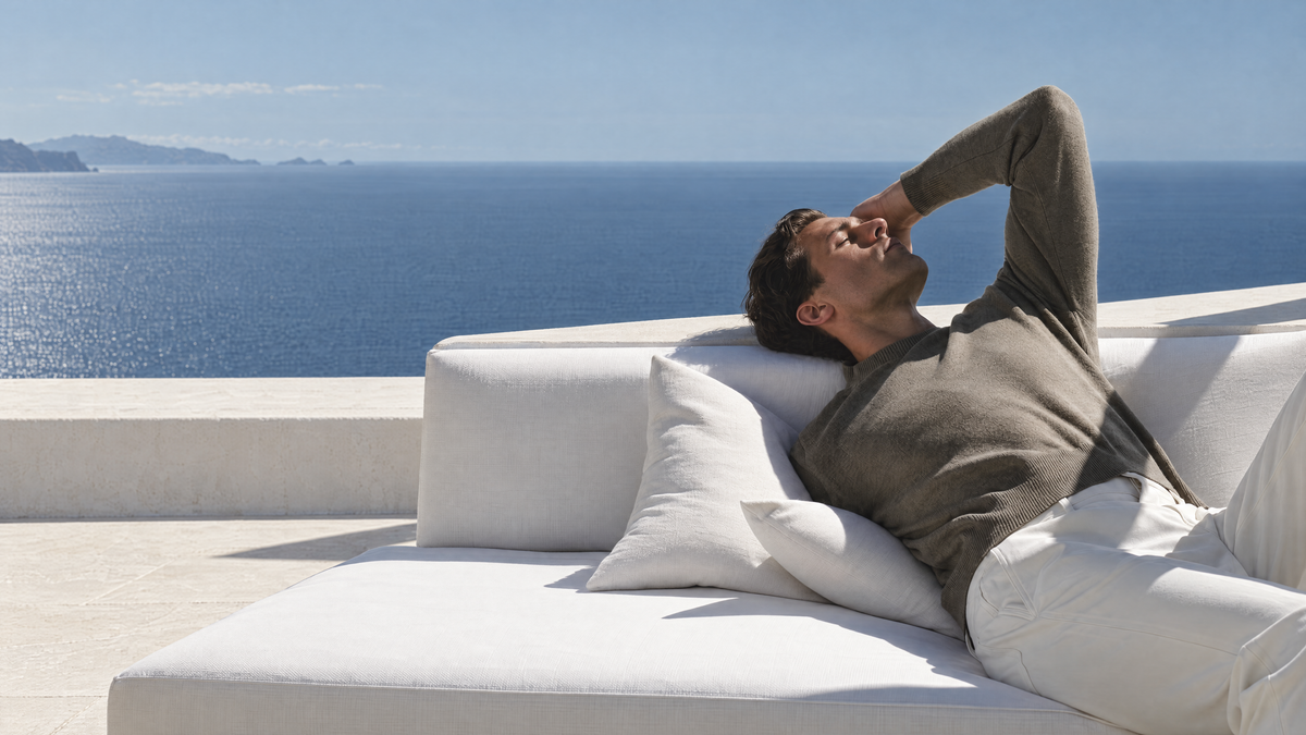 Coastal editorial of a man in taupe knit reclining on an ivory daybed